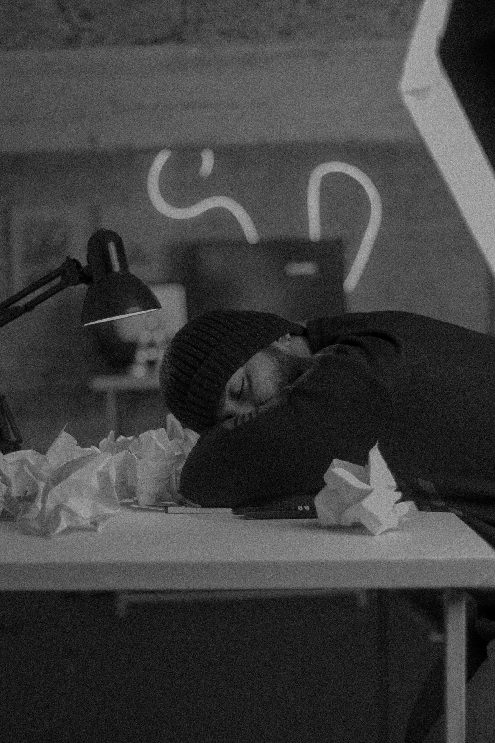 A man in a beanie asleep on a desk surrounded by crumpled papers.